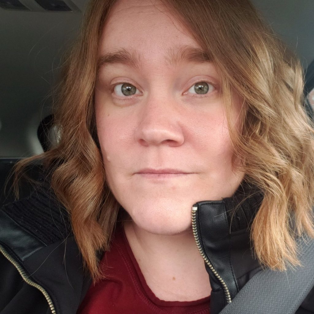 E.K. Johnston wears a black coat and red t-shirt. Behind her is the interior of a car and a seatbelt rests across her shoulder.