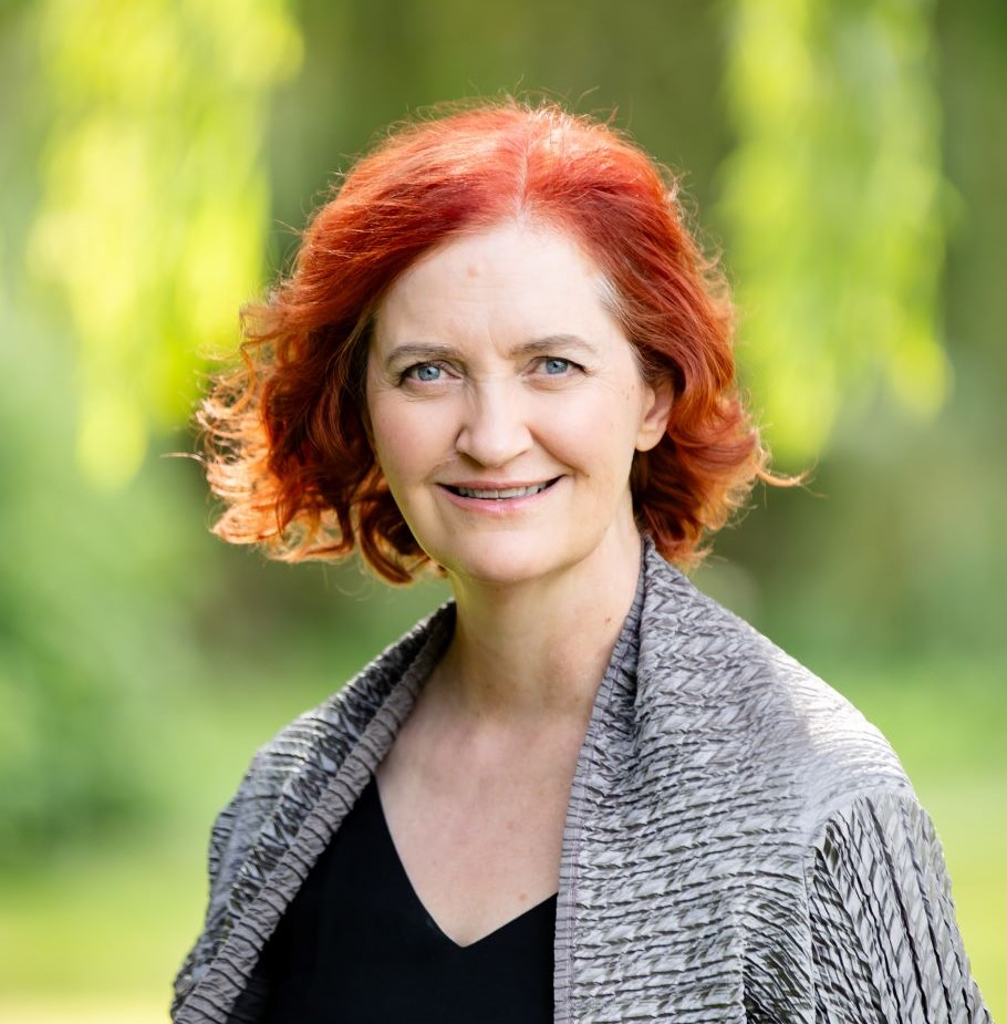 Emma Donoghue wears a gray knit cardigan and black v-neck shirt. Behind her is a blurred background of green foliage.