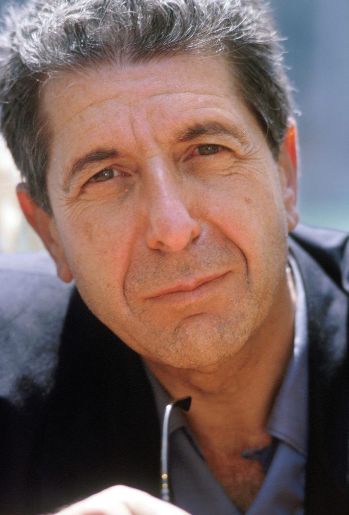 Leonard Cohen wears a black suit. Behind him is a blurred blue and tan background.