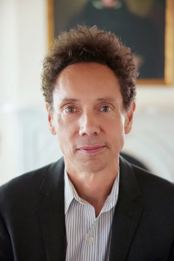 Malcolm Gladwell wears a black suit and gray and white striped shirt. Behind him is a blurred background with a framed painting and white carved fireplace.
