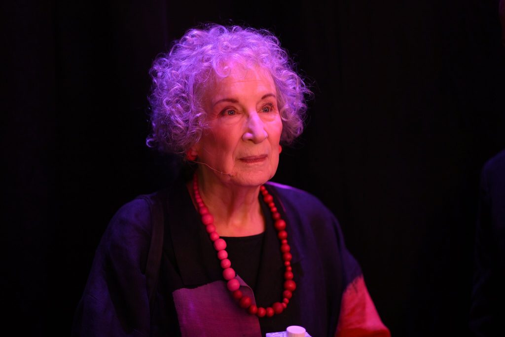 Margaret Atwood wears a blue, black, purple, and red shirt with a pink and red beaded necklace and red beaded earrings. She is in front of a black background with a purple stage light shining on her. A small microphone is attached at her jawline.