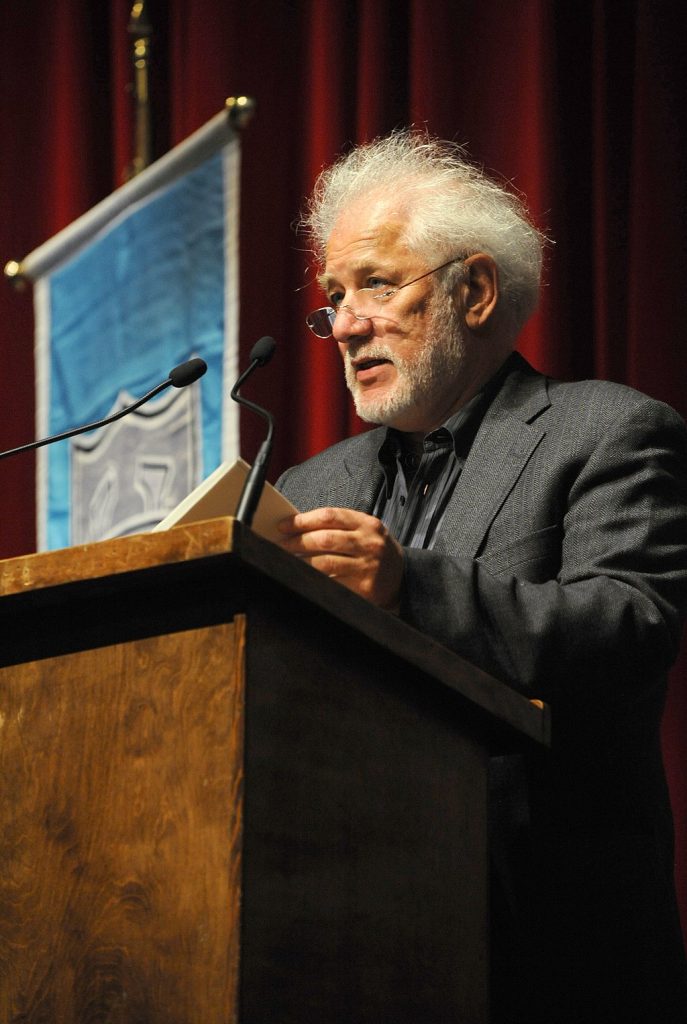 Michael Ondaatje wears a gray suit and rimless glasses. He stands behind a wood podium. Behind him is a dark red curtain and a blue flag with a gray crest.