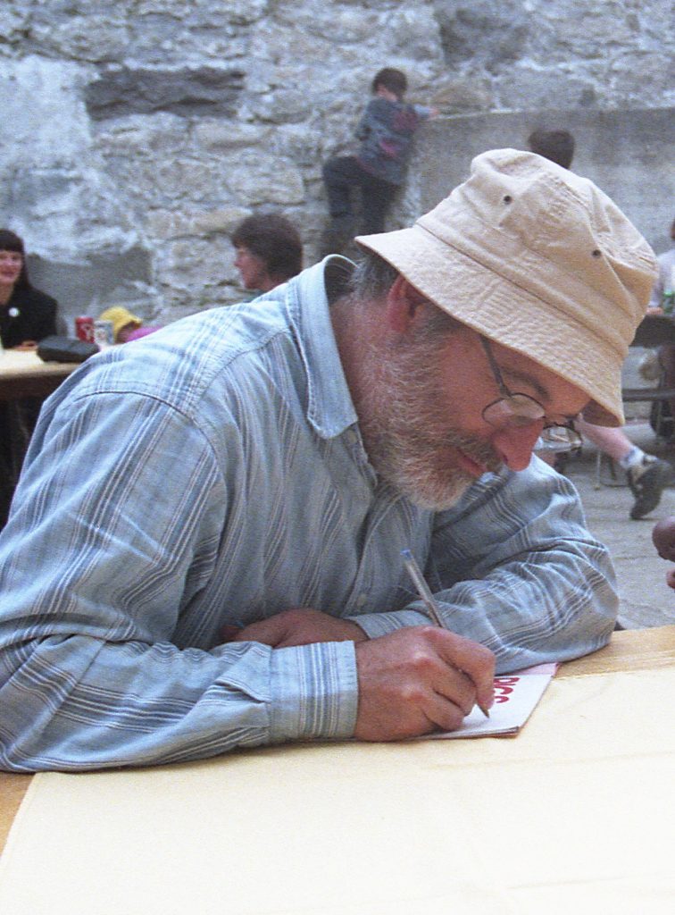 Robert Munsch wears a blue striped shirt, a khaki bucket hat, and black metal glasses. He leans over a wood table and signs a book with a pen. Behind him is a gray stone wall and other people sitting at various tables.