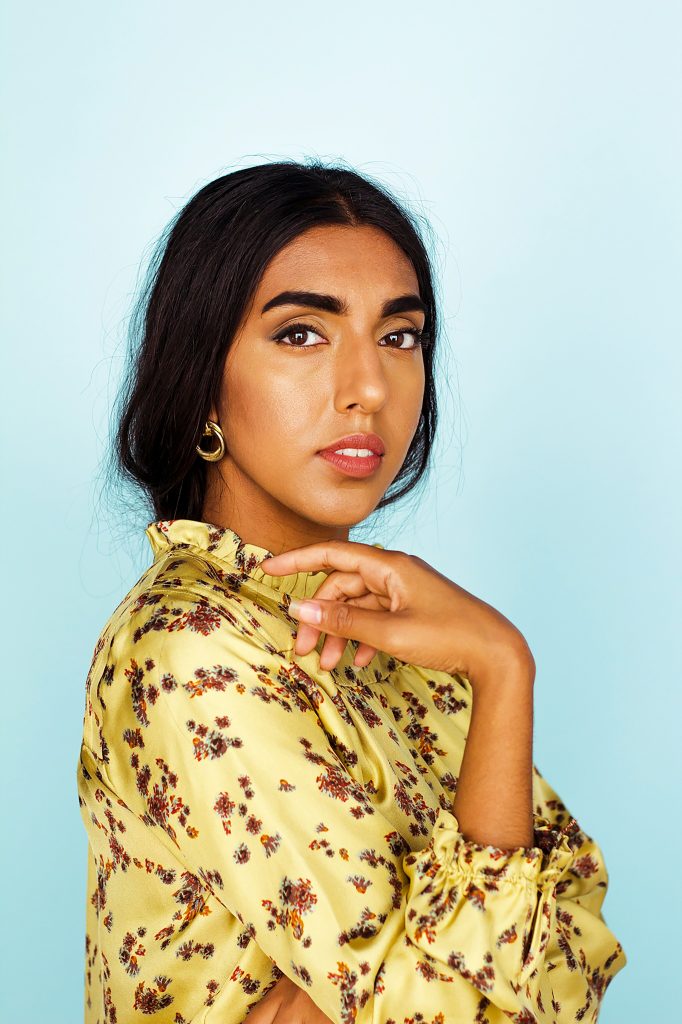 Rupi Kaur wears a yellow floral blouse and gold earrings. Behind her is a pale blue background.
