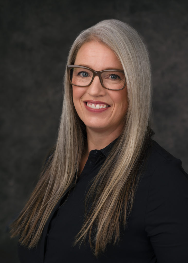 Susan Juby wears a black button down shirt and brown glasses. Behind her is a dark gray background.