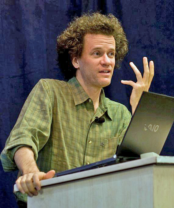 Yann Martel wears a green plaid shirt and stands at a gray podium with a black laptop. Behind him is a dark blue curtain.