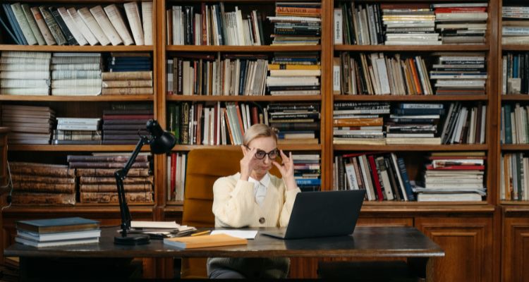 A woman with glasses sitting at a desk in front of bookshelves, looking at a laptop.