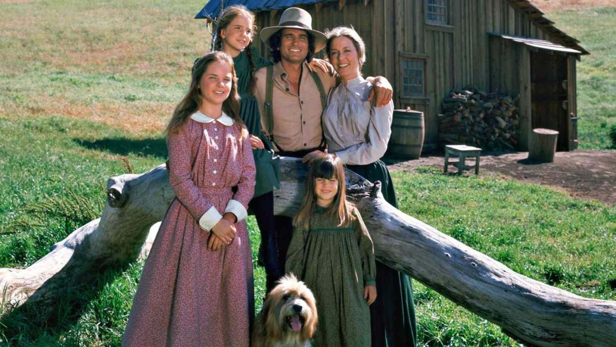 A light-skinned family consisting of father, mother, and three daughters and a shaggy dog pose smiling in front of a rough-hewn wooden home on a prairie.