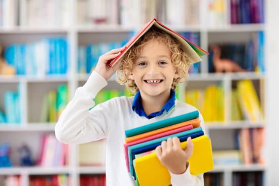 Young smiling boy holds a stack of books and holds an open book on his head.