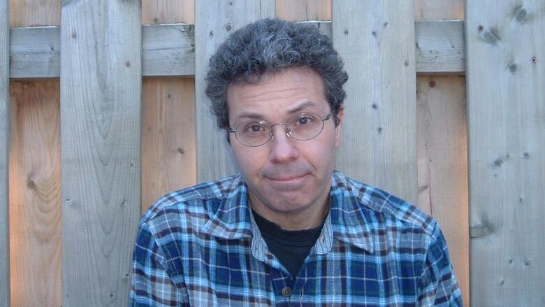A man with curly hair and glasses sits in front of a fence