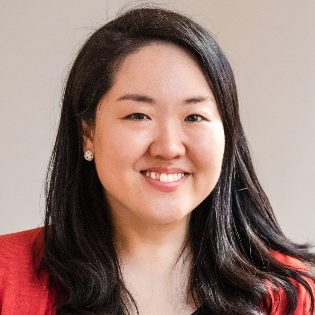 A smiling light-skinned woman with east-Asian facial features and long dark hair wearing a red blouse against a beige background.