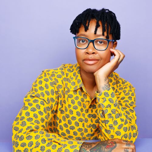 Brittney WIlliams wearing eyeglasses, a yellow patterned button-down shirt. Their tattooed hands show as they lean their chin on their palm against a purple backdrop.