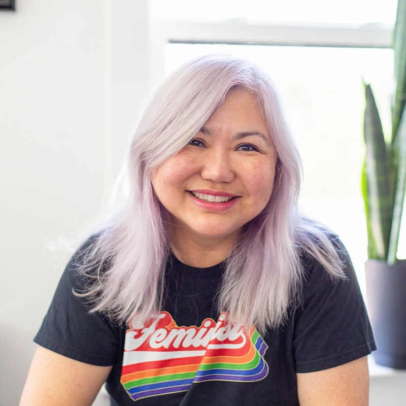 A smiling light-skinned woman with east-Asian facial features and medium-length lavender-colored hair wearing a black t-shirt with the word "Feminist" in rainbow text in front of a white wall and open window with a snake plant on the windowsill.