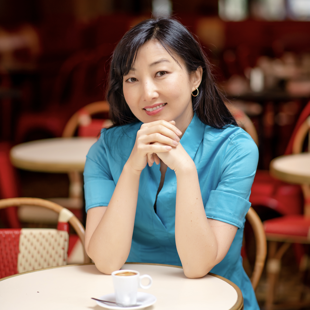 A smiling light-skinned woman with east-Asian facial features and long dark hair wearing a blue blouse is seated with her hands folded and elbows propped against on a cafe table that holds a cup of espresso.