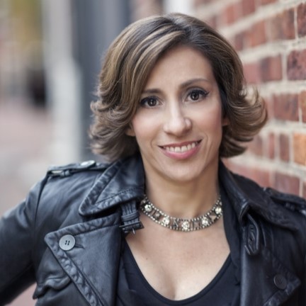 Kami Garcia, wearing a black leather jacket over a black blouse, leans against a brick wall outdoors.