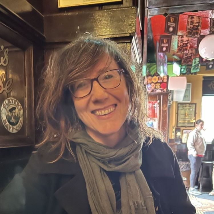 A smiling light-skinned woman with dirty blonde hair and wearing glasses seated in the interior of a pub.
