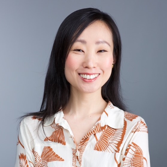 A smiling light-skinned woman with east-Asian facial features and long dark hair wearing a white and orange patterned button-down blouse in front of a gray backdrop.