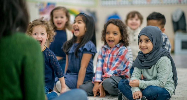 A diverse group of children in skin tone and hair texture sit on a floor, laughing with an adult that sits in front of them.
