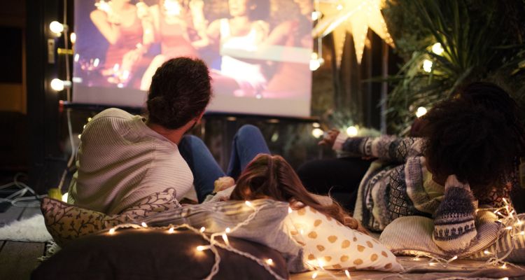 A group of people of various ages, skin tones, and hairstyles lying on pillows and blankets as they watch a movie on a projector framed by fairy lights.