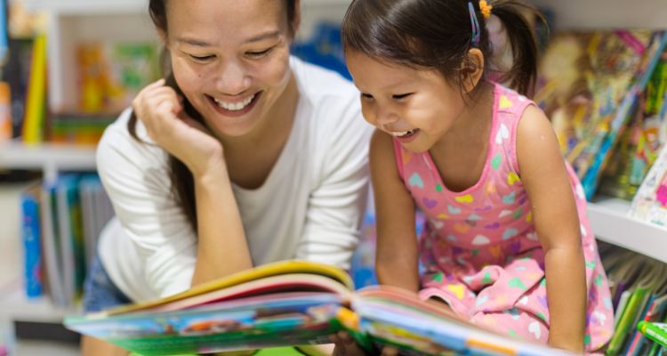 A pale-skinned mother and child with Asian-presenting features and dark hair smile while looking at an open picture book together.