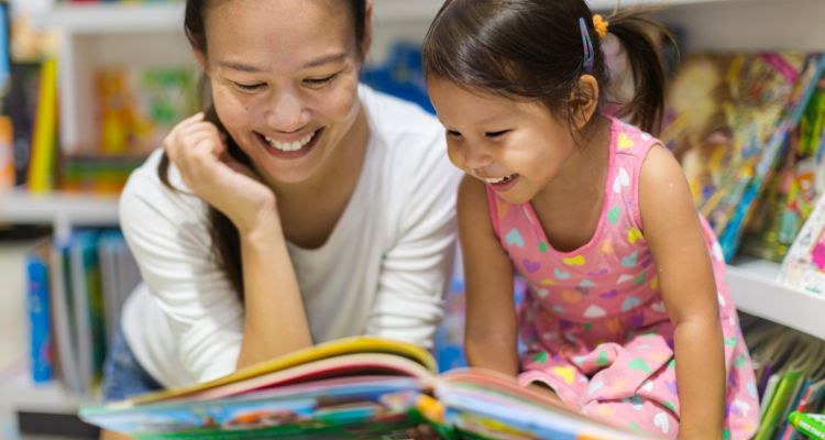 A light-skinned adult and child with east-Asian facial features and dark hair grin down at an open picture book; shelves of more picture books are behind them.