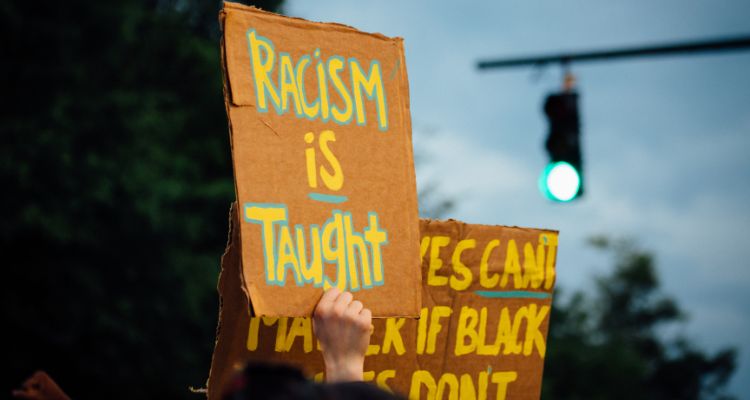 A light-skinned hand holds up a board sign reading "Racism is Taught" in front of other cardboard signs.