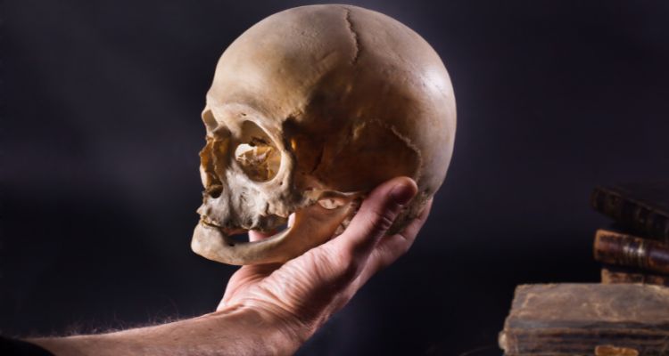 A light-skinned hand holds a skull; antique books are visible in the background.