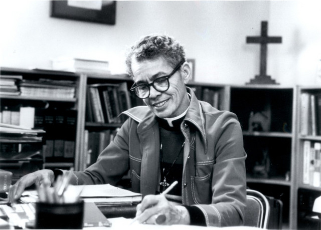 Grayscale photo of Dr. Pauli Murray seated at a desk, writing. Behind her are bookshelves and a framed picture.