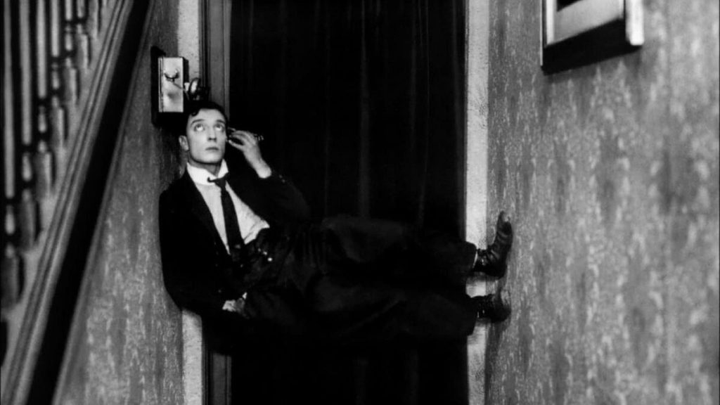 A light-skinned man with short dark hair holds the earpiece of an antique telephone to his ear while balancing horizontally across a narrow hallway by his shoulder and feet.