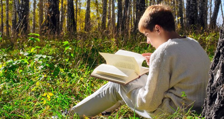 A light-skinned youth with short brown hair sits in a forested area against a tree trunk reading an open book.