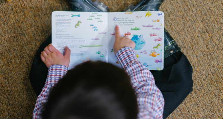 A small child with dark hair is viewed from above as they sit on the ground in galoshes, holding a picture book in their lap and point at the words.