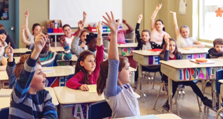 Older elementary-aged children sit at desks in a classroom, nearly all with arms raised to answer a question.