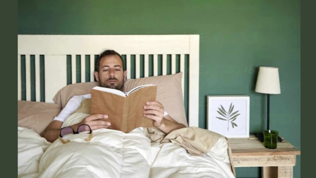 A man lies in his bed reading a book, which is rested atop his chest. On his bedside sits a lamp and decorative frame. The walls around the room are green and compliment the cream coloured sheets and bedframe.