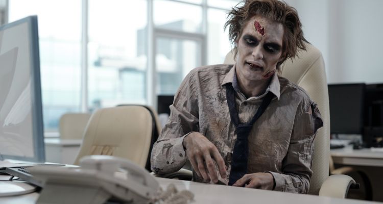 A zombie is wearing a button-up shirt and a tie. He sits at a desk with a computer and a phone.