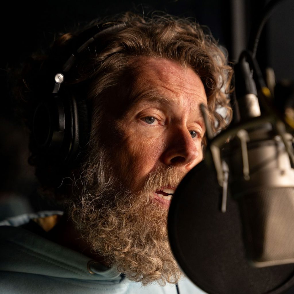 A close-up of a man with tan skin, hazel eyes, and curly reddish-brown hair as well as a beard. He is speaking into a large microphone, and is wearing a light blue sweatshirt and dark headphones.