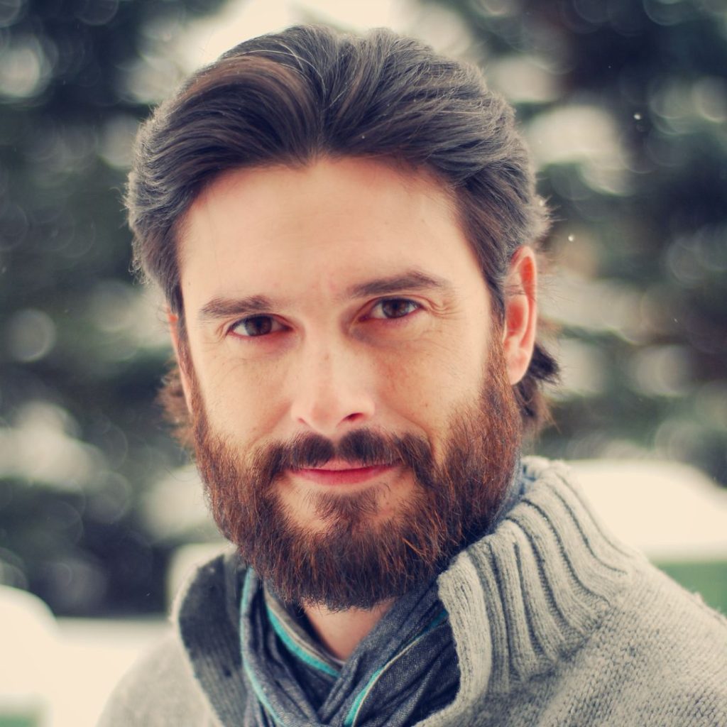 A headshot of a man with light skin, dark eyes, and dark brown hair as well as a trimmed beard. He is wearing a gray sweater and blue-gray scarf and is smiling slightly in front of a blurred snowy background.