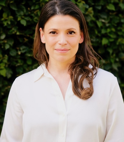 A woman with light skin, dark eyes, and dark brown medium-length hair that lightens to regular brown as it extends. She is wearing an off-white blouse, is smiling slightly, and is standing in front of a leafy background.