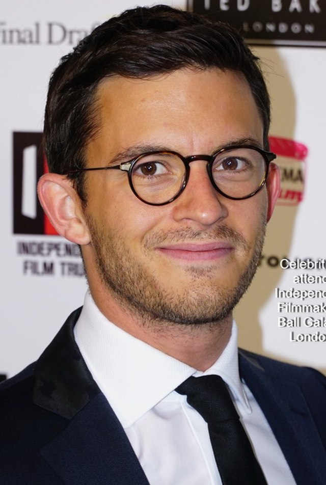 A photo of Jonathan Bailey in 2015 at an event. He's dressed in a black suit, wearing glasses, and smiling.