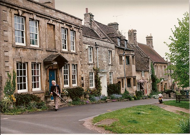 A picture of Bailey's hometown of Oxfordshire. Old brick, English buildings in a row along a paved street. An old man walks past a front door with a cane.