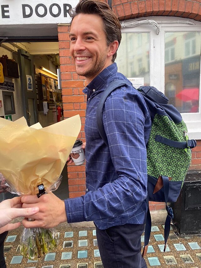 A picture of Bailey of a fan handing him flowers. He's smiling, dressed in a blue plaid button up wearing a green and blue pattern backpack holding coffee.