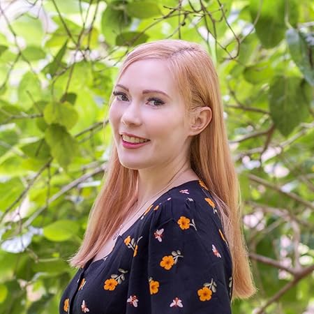 A woman with long blonde hair wears a dark blue shirt with flowers and stands in front of green, leafy plants.