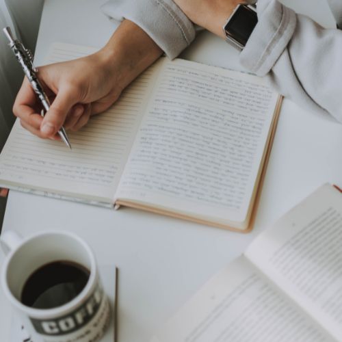 Someone is hovering a metal pen over a journal that has writing on the left page and a few lines done on the other. In fron tof them sits a cup of coffee anf a book that is open in the corner.