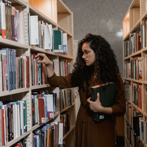 A woman with round glasses holds a big green book in her left hand as she browses a bookshelf to her right.