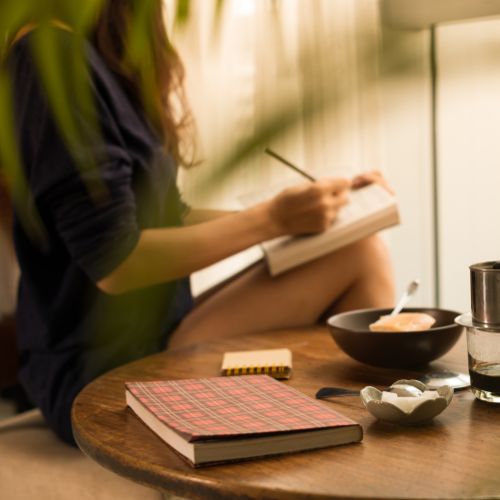 A table sits in front of the camera with a red plaid journal, two bowls, and a spoon next to coffee. Sitting away from the table is a woman sitting up in a blue shirt writing in a journal. leaves from a plant slightly shade her.