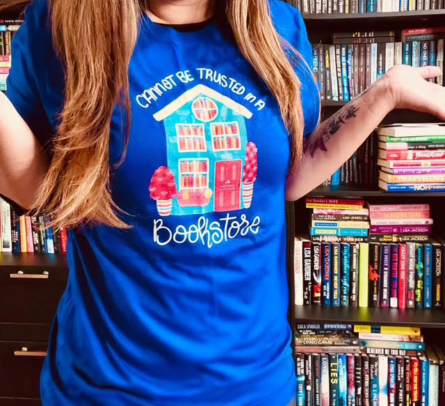 Amy stands in her home-library wearing a bright blue T-Shirt. The shirt has a photo of a light blue building with a red door and four windows. The windows are filled with books. Text on the T-Shirt reads: Cannot Be Trusted In A Bookstore.