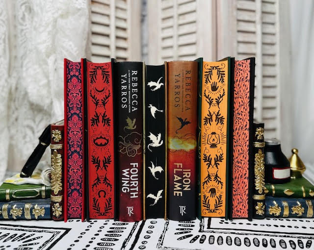 A set of vertically stacked editions of The Empyrian book series by Rebecca Yarros. The spines of Fourth Wing and Iron Flame are visible while the rest are displayed with their sprayed edges facing forward.