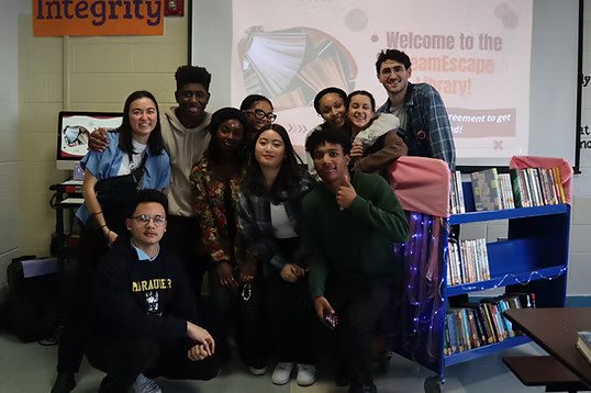 Group of high school students smiling in front of a display welcoming visitors to the DreamEscape Library, with bookshelves and decorations in the background..