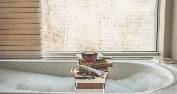 A grainy image of a bathtub full of bubbles beneath a window with a sheer covering. A balanced tray rests across the tub with a glass of red wine, and a book.