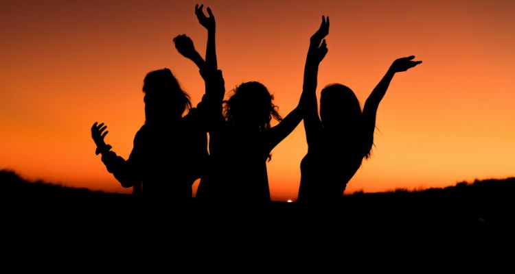 Three figures stand in front of a sunset with their arms raised up.