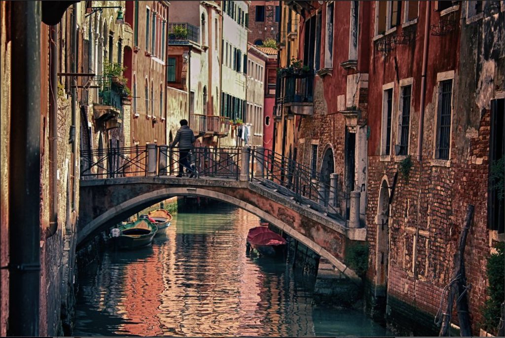 Person walking on a small bridge over a small canal. The bridge spans rows of old brick buildings.